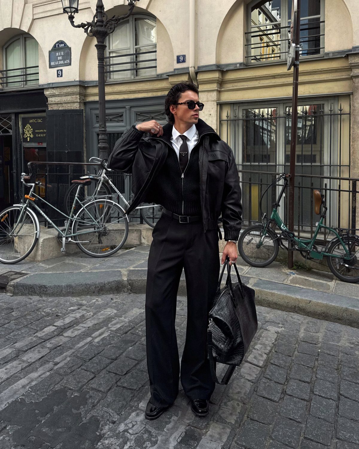 Man in a black jacket, white shirt, and black trousers standing on a cobblestone street.
