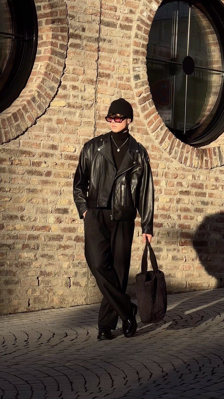 A person in a black leather jacket and turtleneck posing against a brick wall.