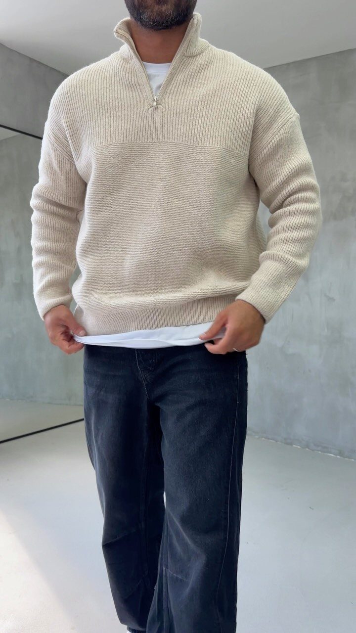 Man wearing a beige ribbed knit quarter-zip sweater and black jeans in a grey room.