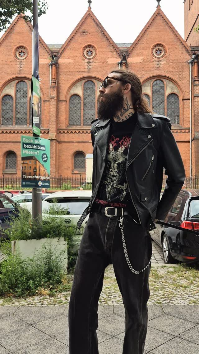 Man with a beard wearing a black leather biker jacket and black jeans standing outdoors.