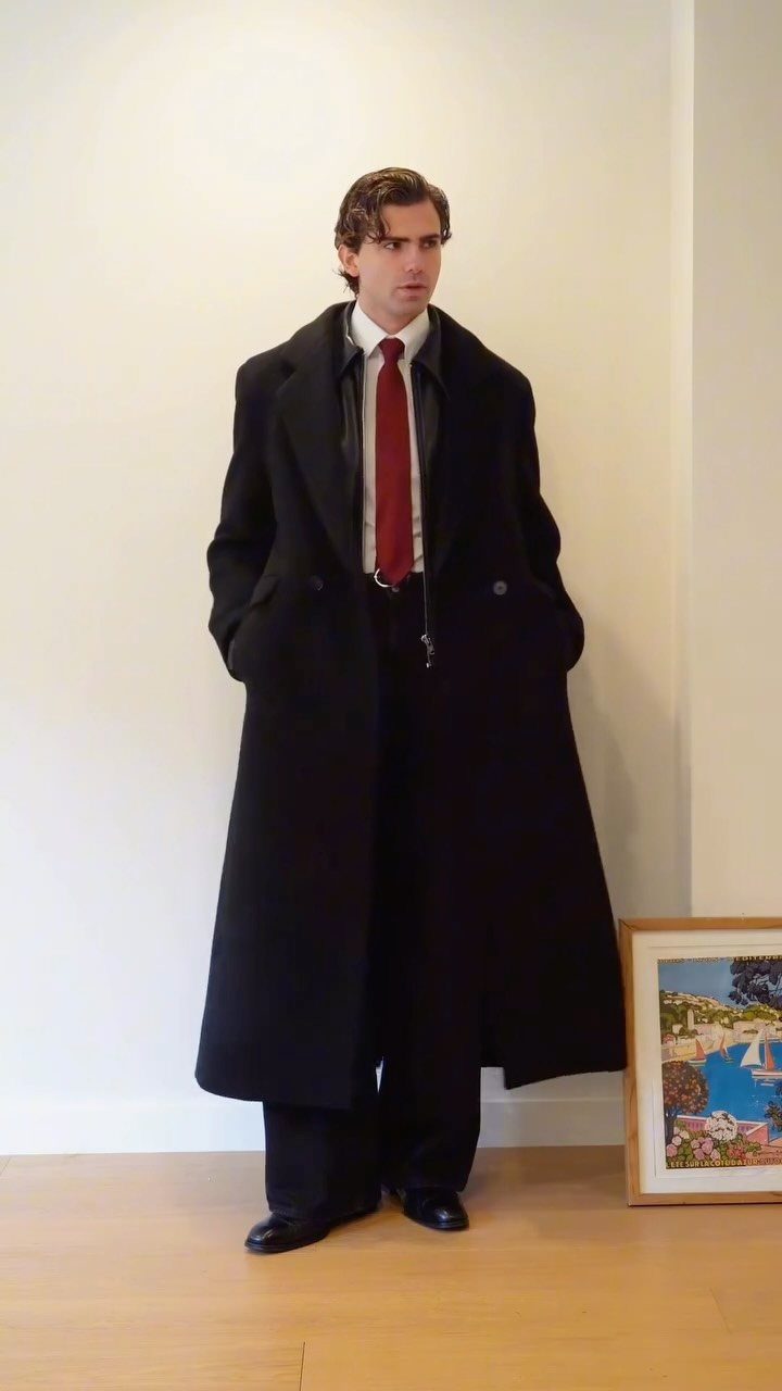 A man wearing a long black coat, white shirt, red tie, and black trousers standing indoors.