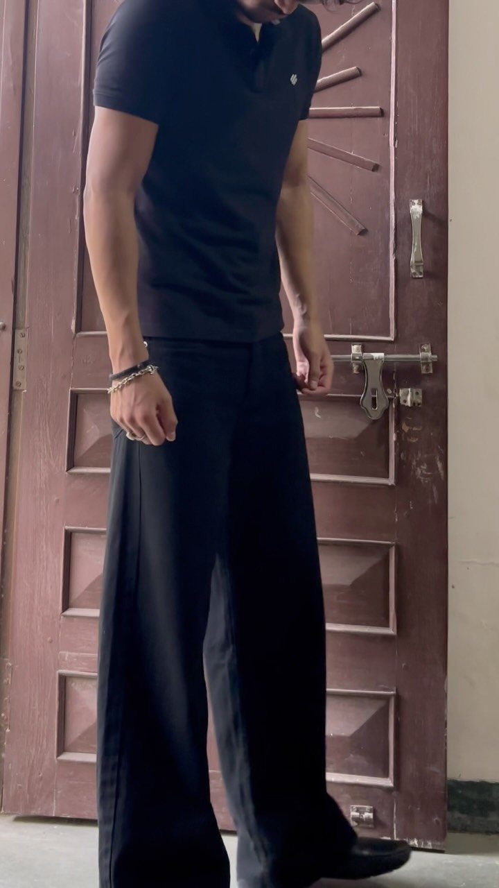 A person wearing a black polo shirt and wide-leg black trousers standing by a door.