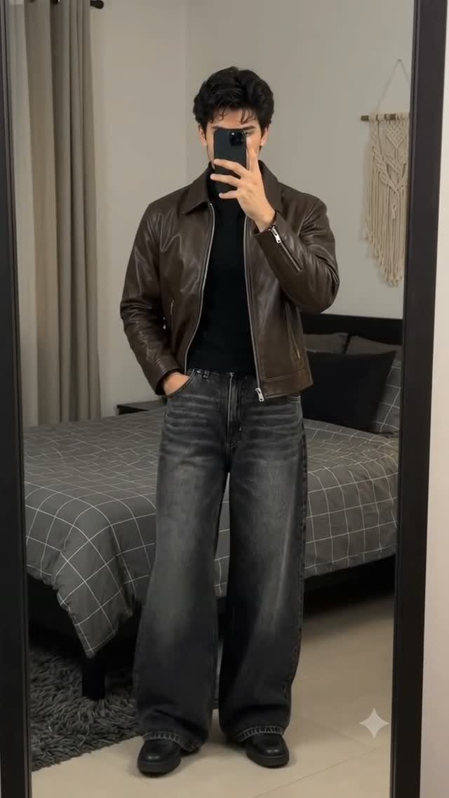 Man in a brown leather jacket, black turtleneck, and wide-leg jeans posing for a mirror selfie.