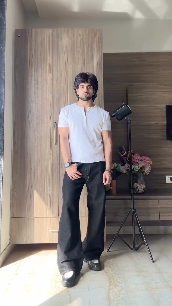 A man posing in a white shirt and wide-leg black jeans indoors.