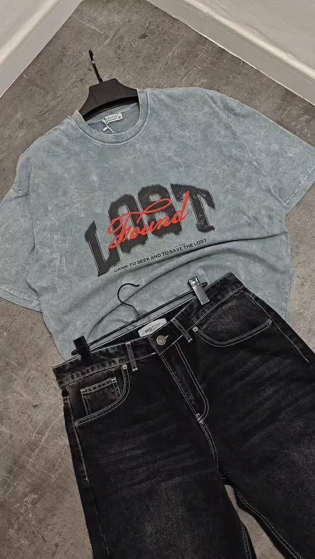 Light blue graphic t-shirt and black acid-wash jeans laid flat on a grey floor.