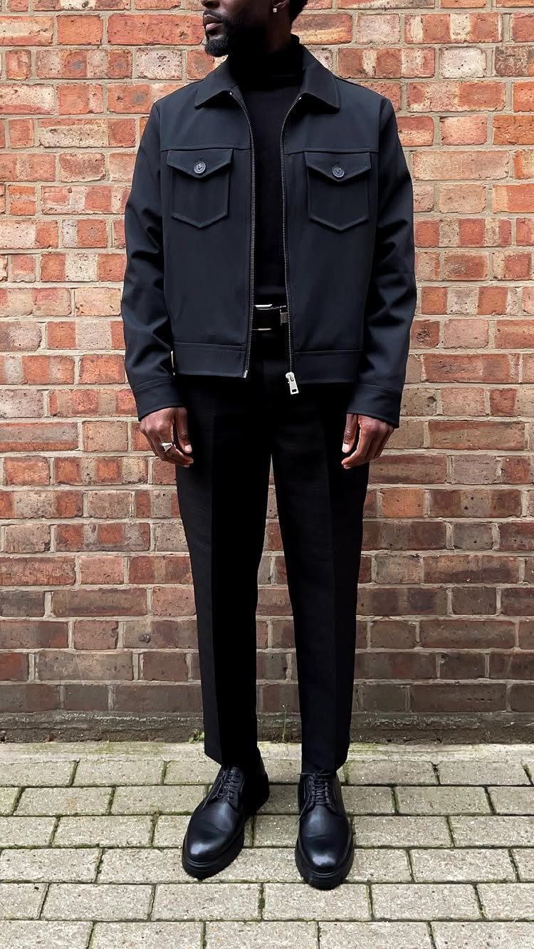 Man wearing an all-black outfit with a jacket, turtleneck, trousers, and leather shoes against a brick wall.