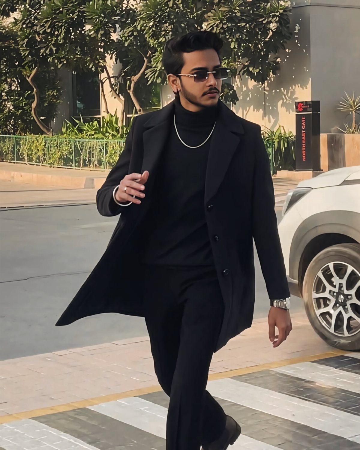 A man in an all-black outfit including a long black overcoat walking on a street.