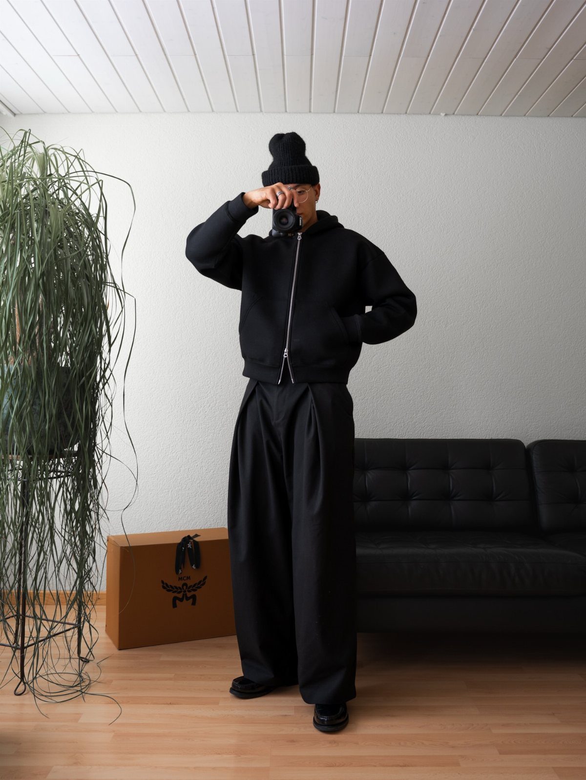 Man in an all-black outfit including a beanie, hoodie, and wide trousers standing in a room.
