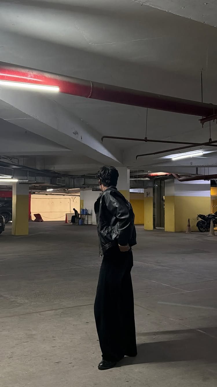 A person wearing a black leather jacket and wide-leg black trousers standing in a parking garage.