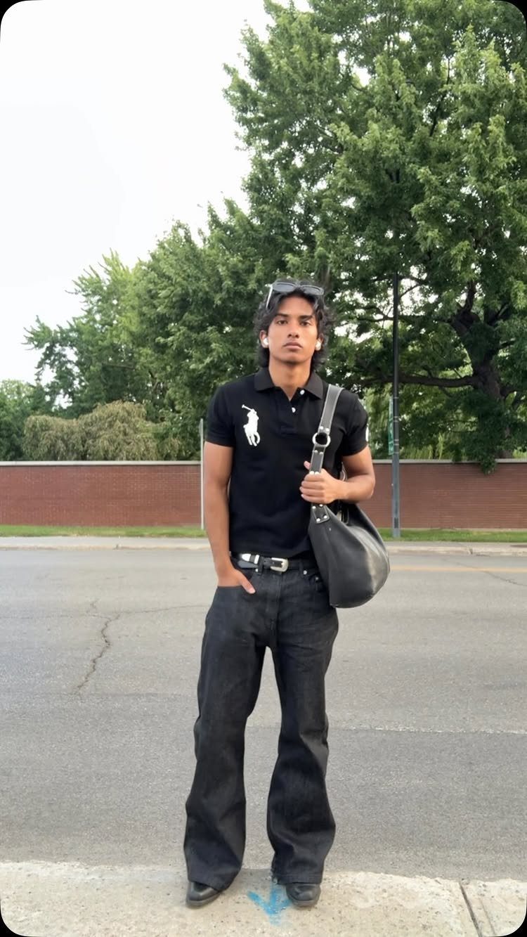 A man wearing a black polo shirt and black flared jeans standing on a sidewalk.