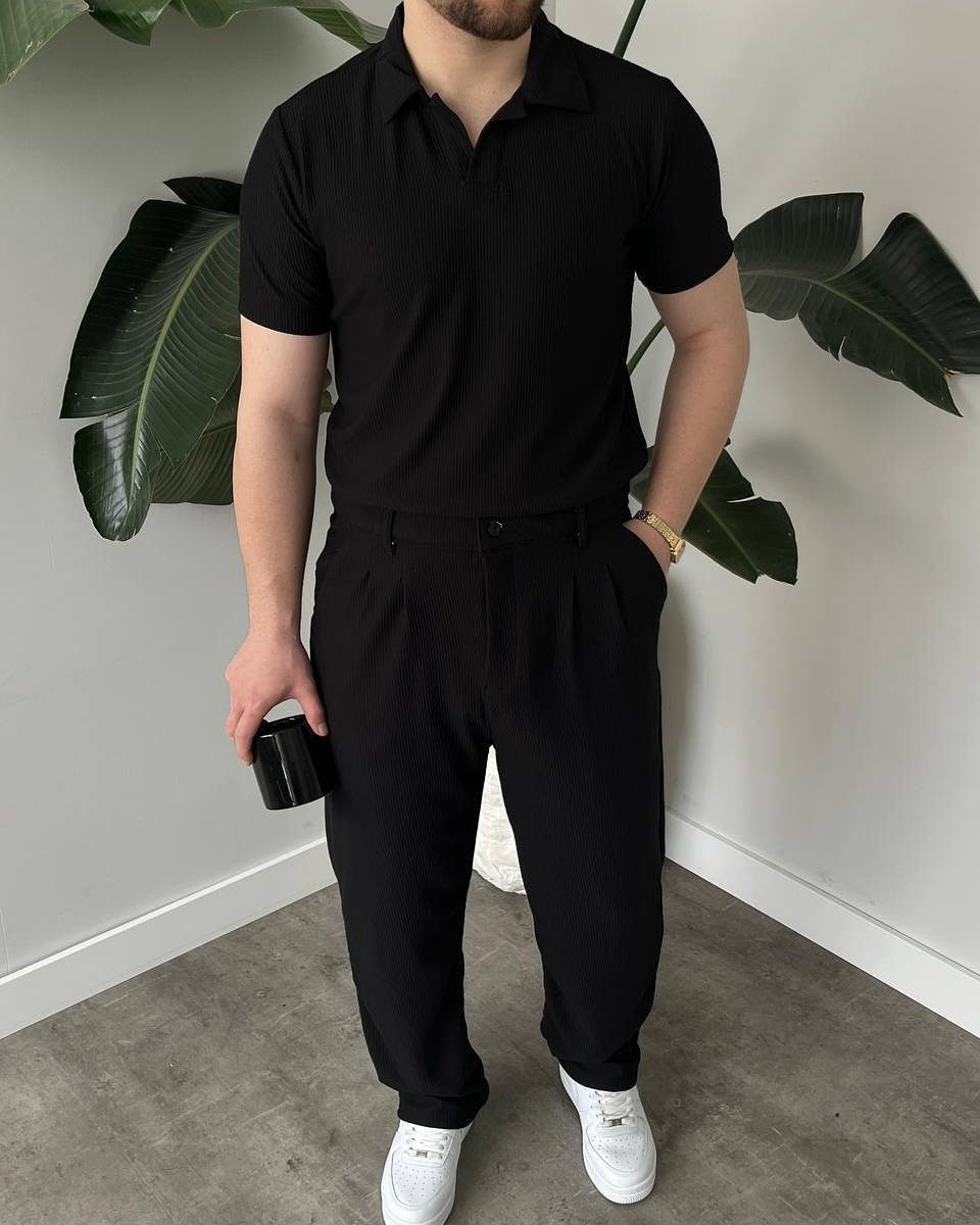 A man wearing a black polo shirt and black trousers with white sneakers standing by tropical leaves.