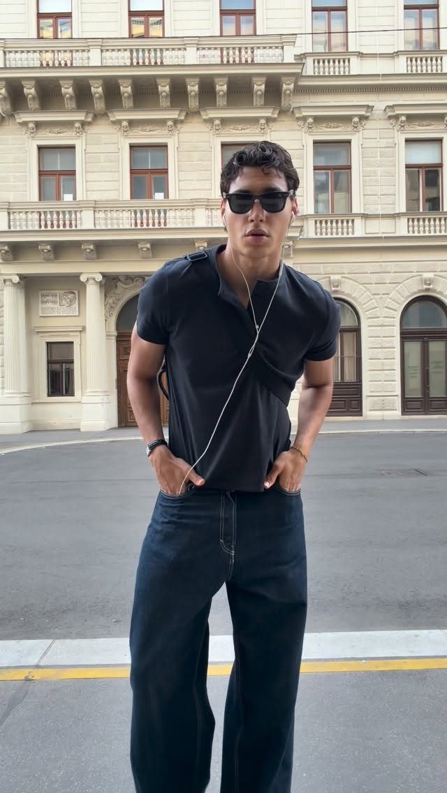 A man in a black polo shirt and dark jeans posing in front of a classical building.