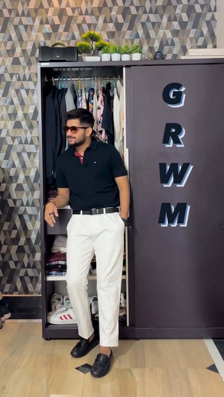 Man wearing a black polo shirt and white trousers standing in front of a wardrobe.