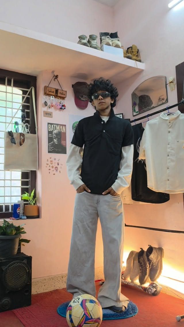 A young man posing in a black polo shirt, white long-sleeve undershirt, and grey wide-leg trousers.