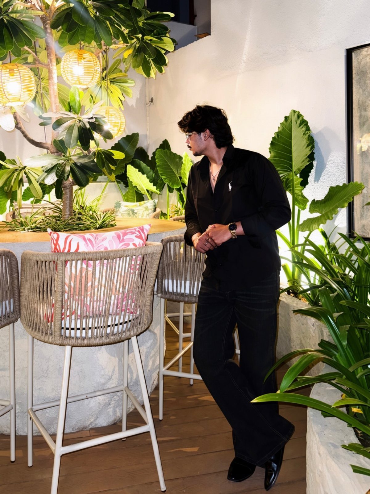 A man wearing an all-black outfit standing in a lush outdoor patio setting.