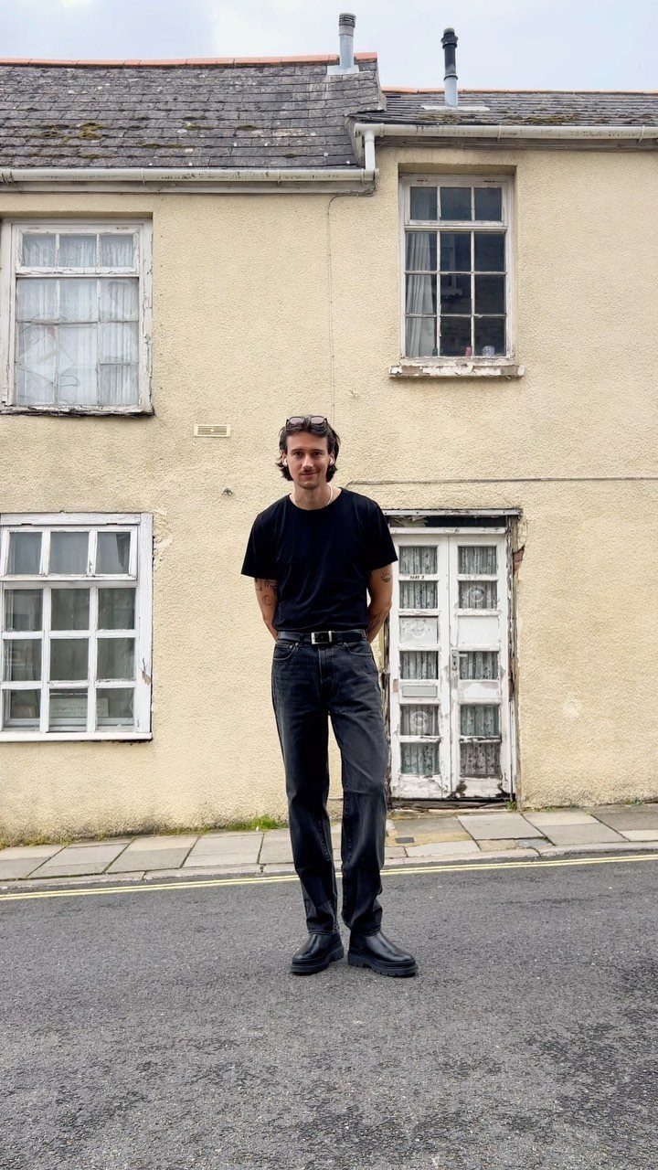 Man wearing a black t-shirt and dark jeans standing on a street.