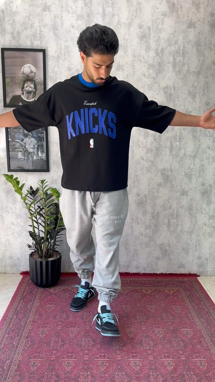 Man wearing an oversized black Knicks t-shirt and grey joggers standing on a red rug.