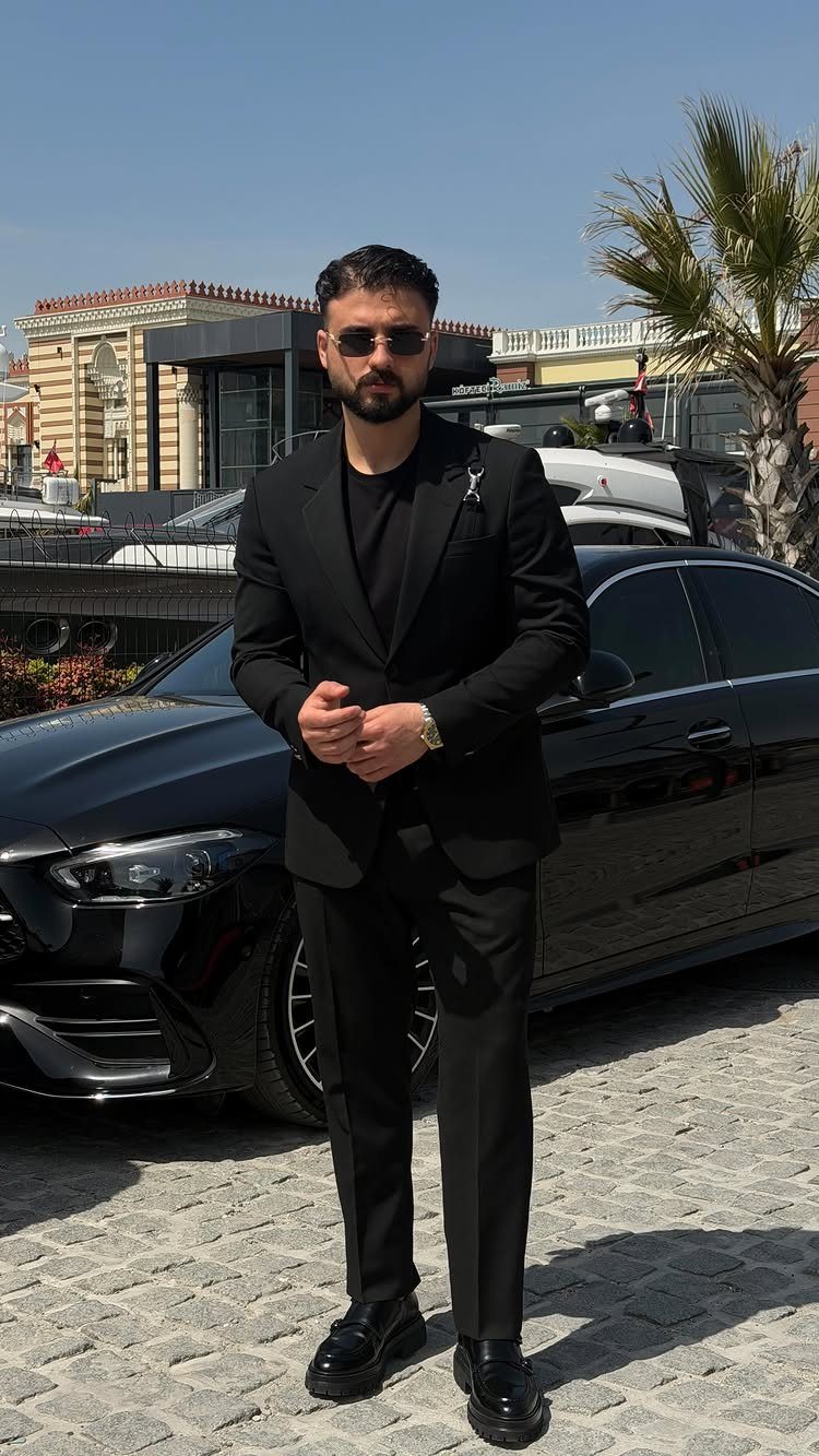 Man in a black suit and t-shirt standing in front of a black car.