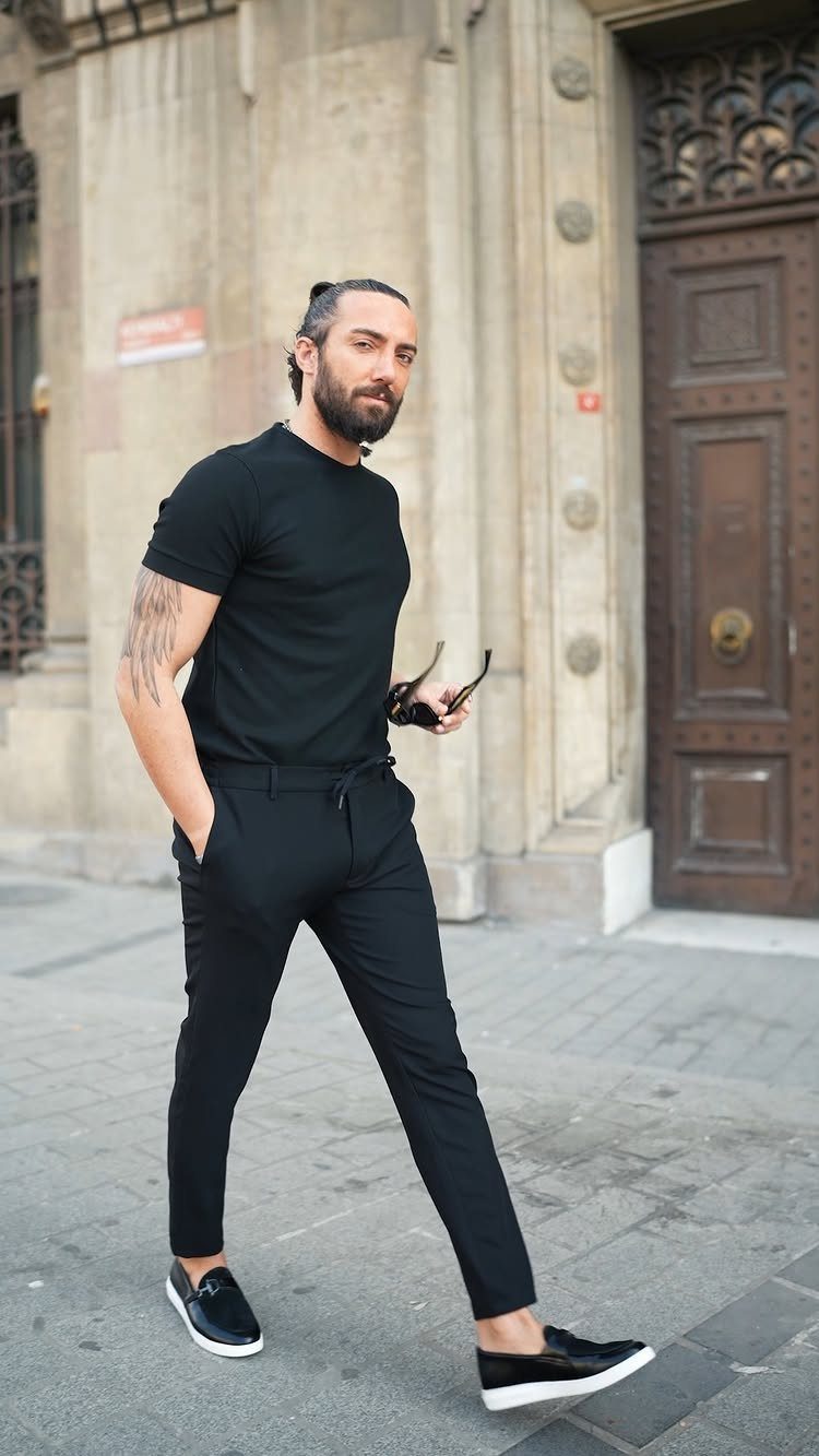 Man walking in a city wearing a black t-shirt and black trousers with slip-on shoes.