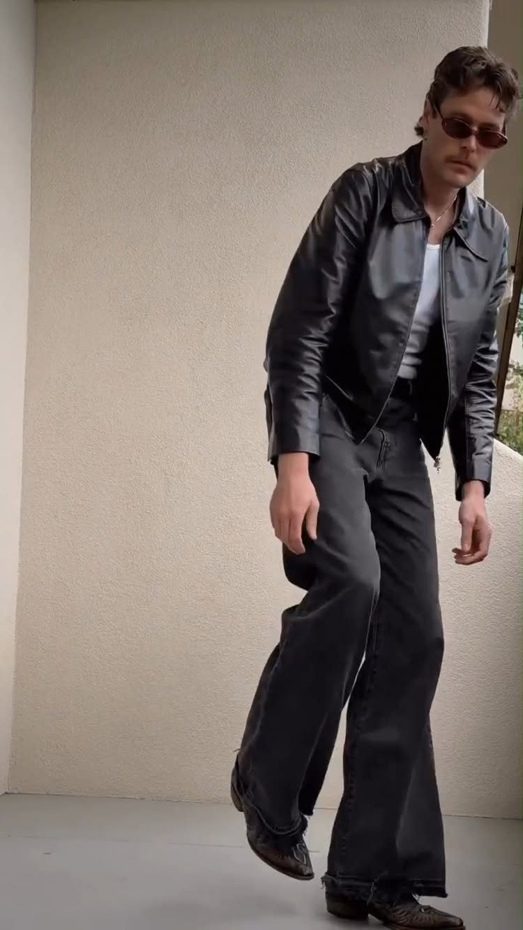 Man in a black leather jacket, white t-shirt, and black wide-leg jeans standing outdoors.