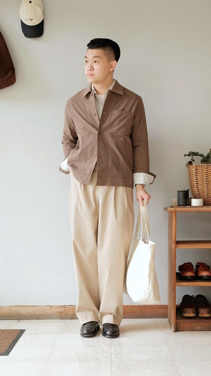 Man wearing a brown jacket, beige trousers, and black loafers while holding a white tote bag.