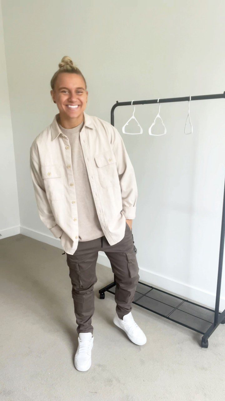 Smiling person wearing a beige button-down shirt, tan t-shirt, brown cargo pants, and white sneakers.