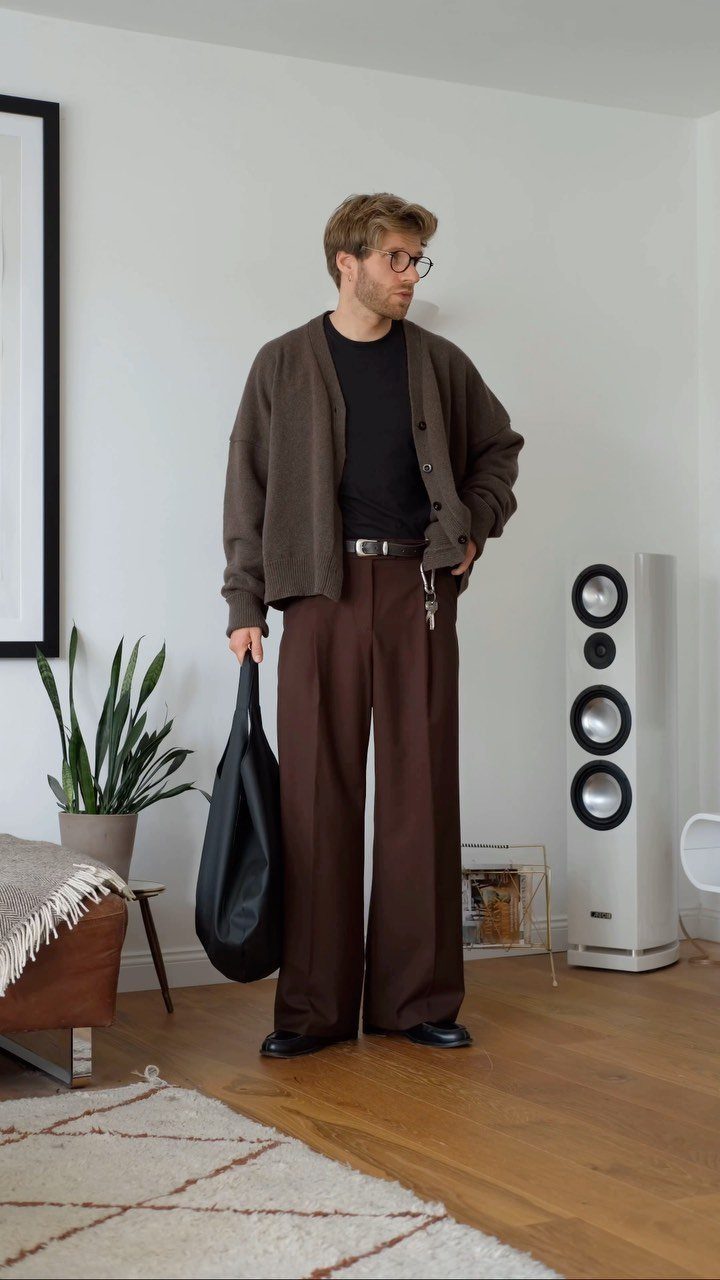 Man standing in a room wearing a brown cardigan, black t-shirt, and wide-leg brown trousers.