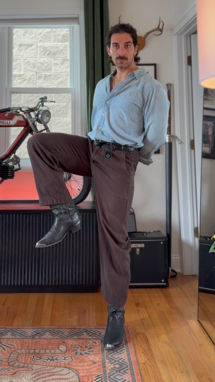 Man posing in a light blue chambray shirt and brown corduroy pants with black boots.