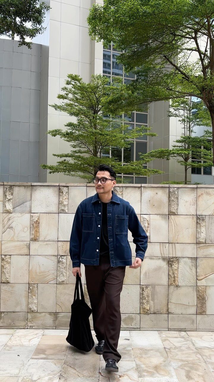 A man walking in a dark blue denim jacket and brown pants while carrying a black bag.