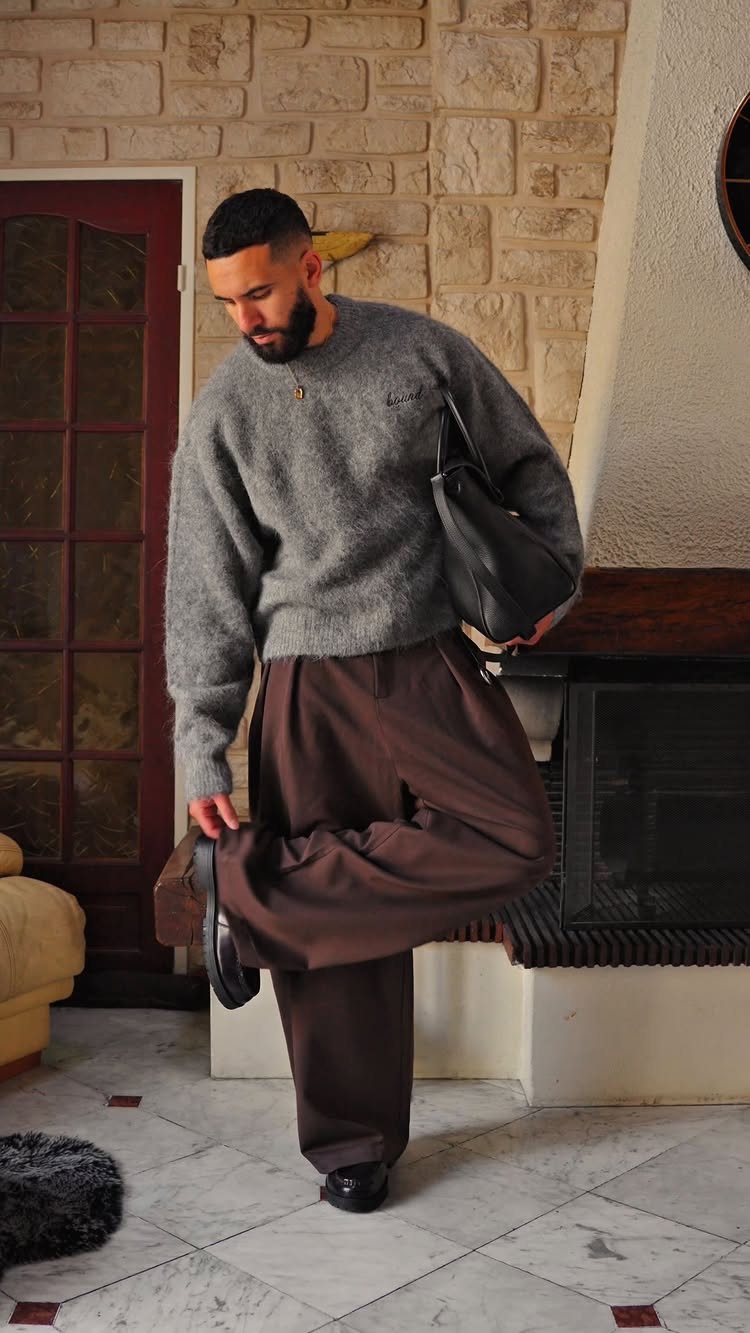 Man in a grey sweater and wide-leg brown trousers standing near a stone fireplace.
