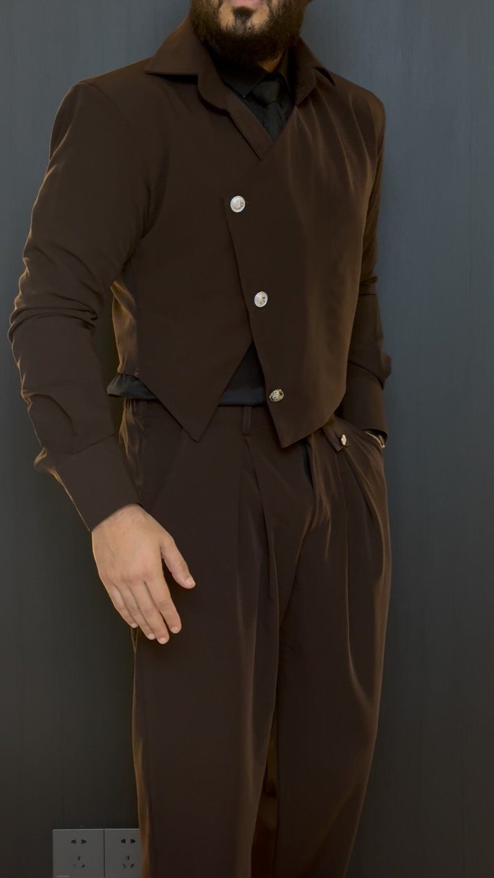 Man wearing a dark brown asymmetrical jacket and matching brown trousers against a grey wall.