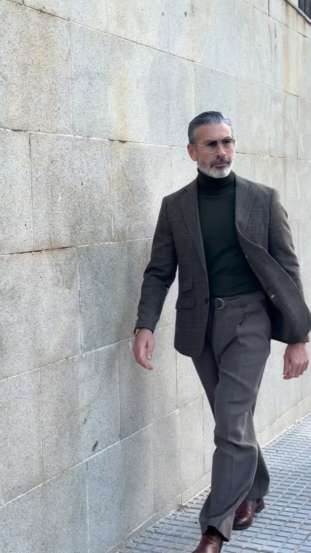 Man walking in a brown blazer, dark green turtleneck, and brown trousers against a stone wall.