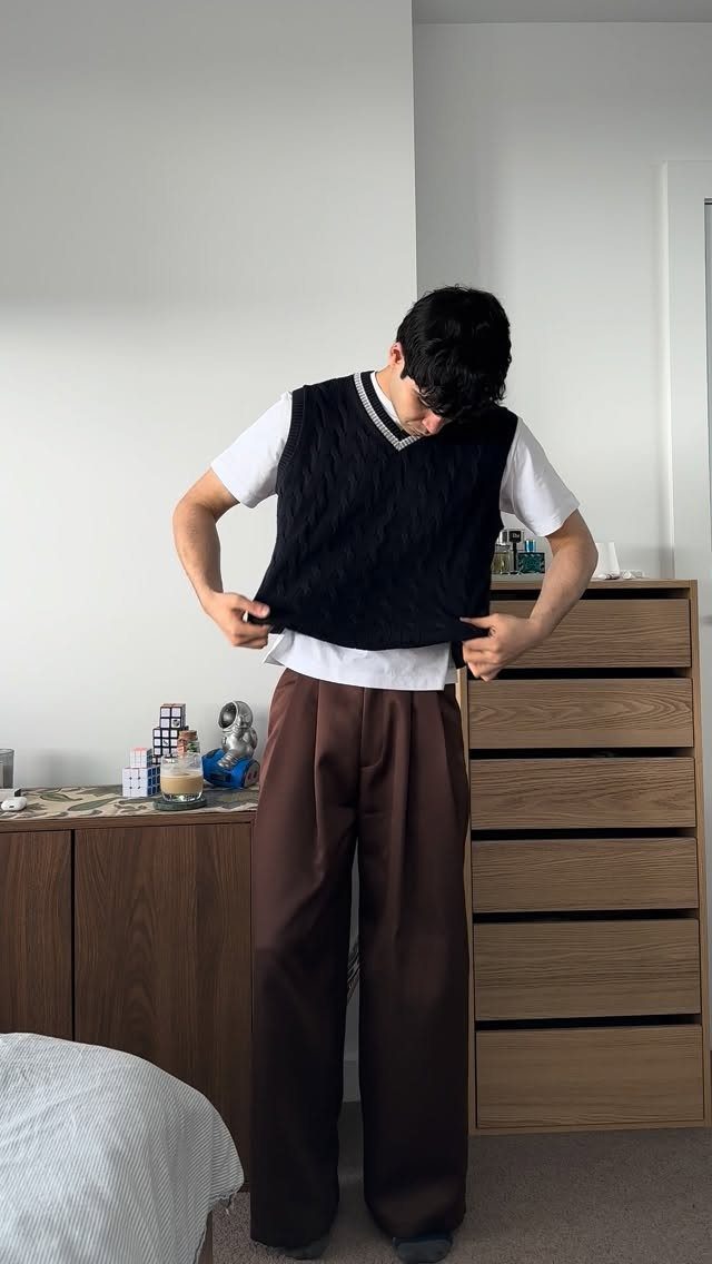 Man wearing a dark sweater vest over a white t-shirt and brown wide-leg pants.