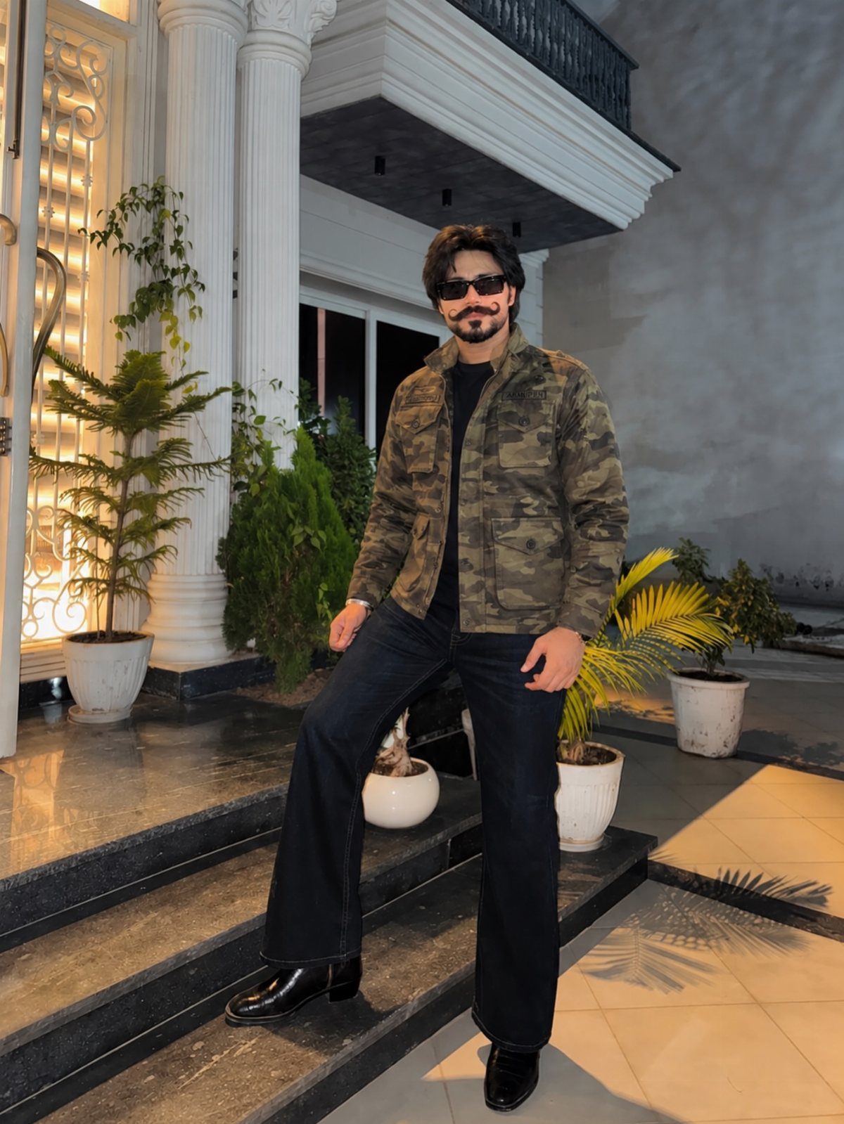Man in a camouflage jacket, black shirt, black jeans, and black boots posing outdoors.