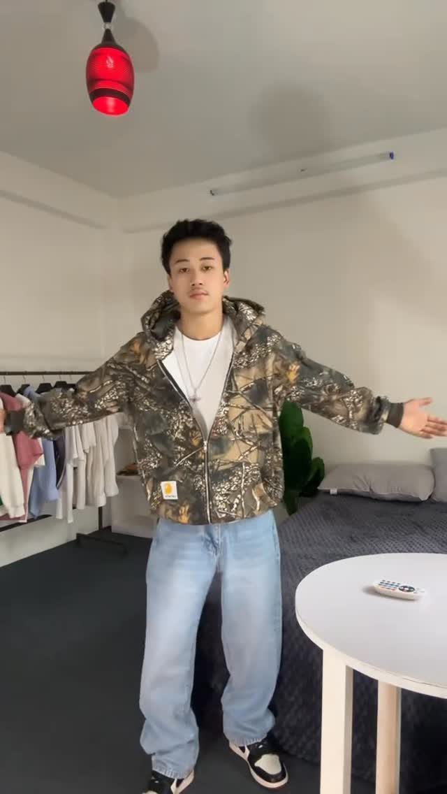 A man posing in a camouflage jacket, white t-shirt, and light blue denim jeans.