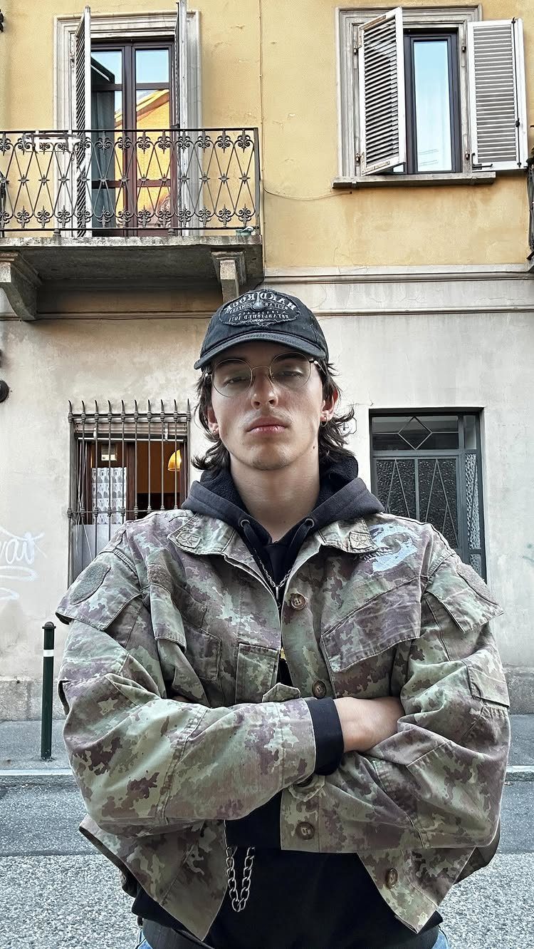 A young man wearing a camouflage jacket, black hoodie, and baseball cap poses outdoors.