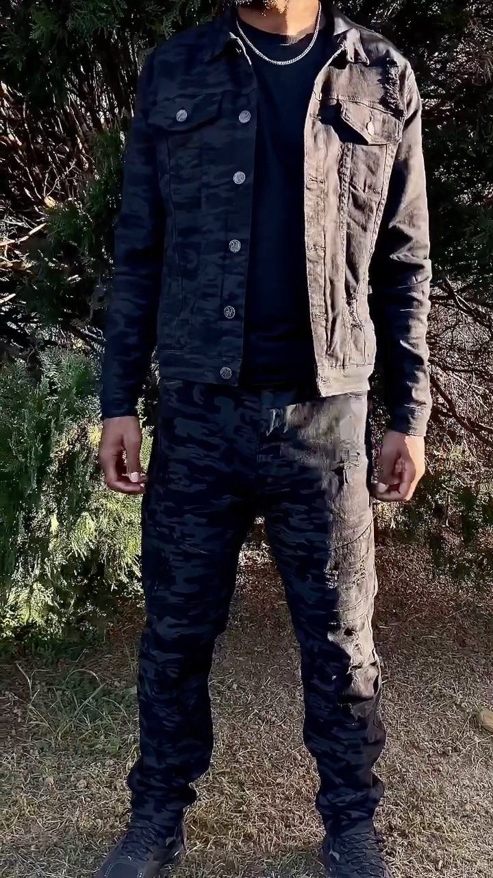 A person standing outdoors wearing a dark camouflage jacket, black t-shirt, and matching camouflage pants.
