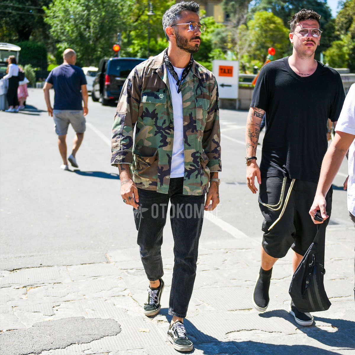 Man walking outdoors wearing a camouflage jacket, white t-shirt, black pants, and sneakers.