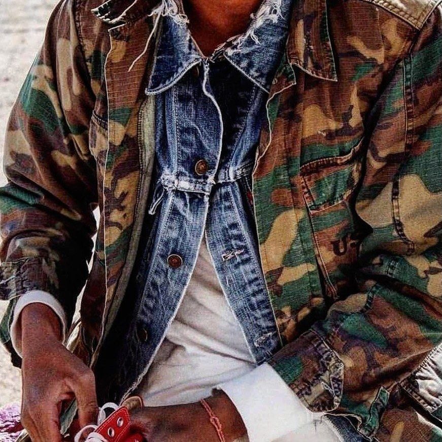 A person wearing a camouflage jacket layered over a blue denim shirt and white top.