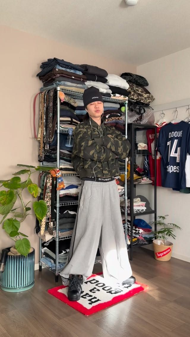 A person wearing a camouflage jacket, black beanie, and wide-leg grey pants standing in a room.