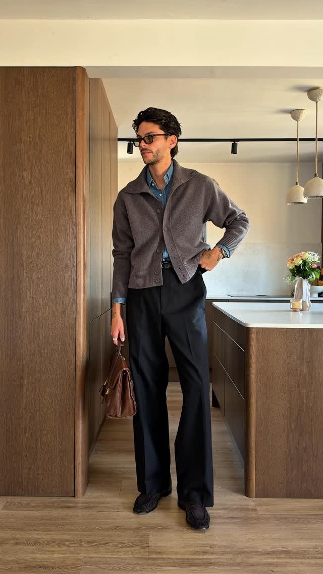 Man in a grey cardigan and wide-leg trousers holding a leather briefcase in a kitchen.