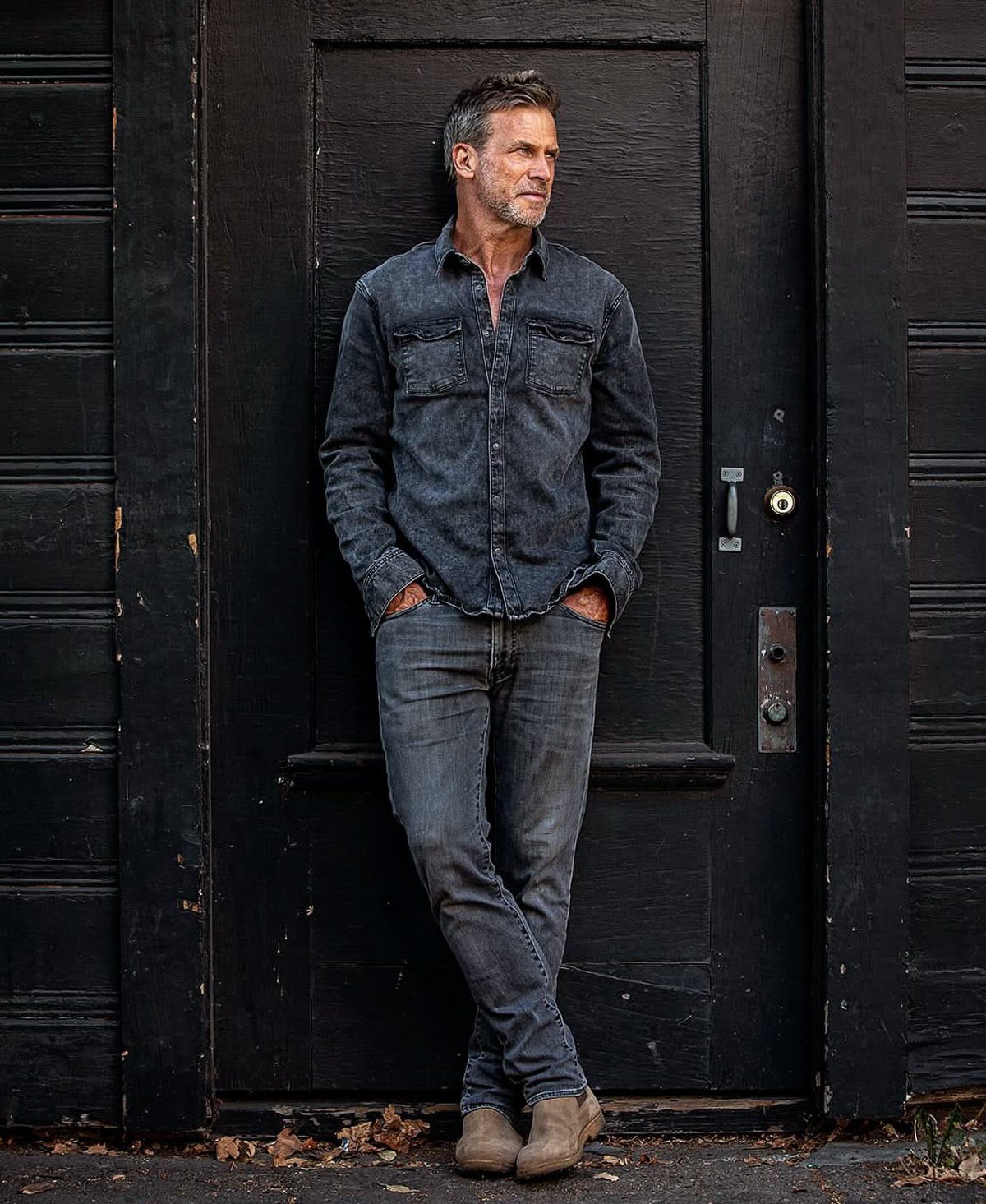 A man in a dark denim shirt and jeans leaning against a black wooden door.