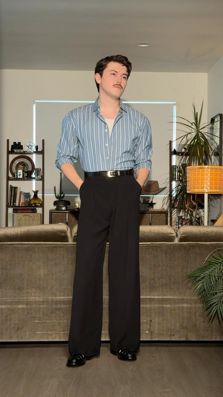 Man with a mustache wearing a striped shirt and wide-leg black trousers standing in a room.