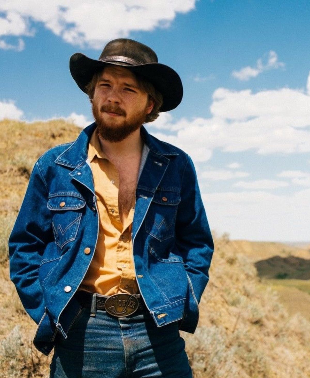 Man in western attire wearing a cowboy hat, denim jacket, and large belt buckle outdoors.
