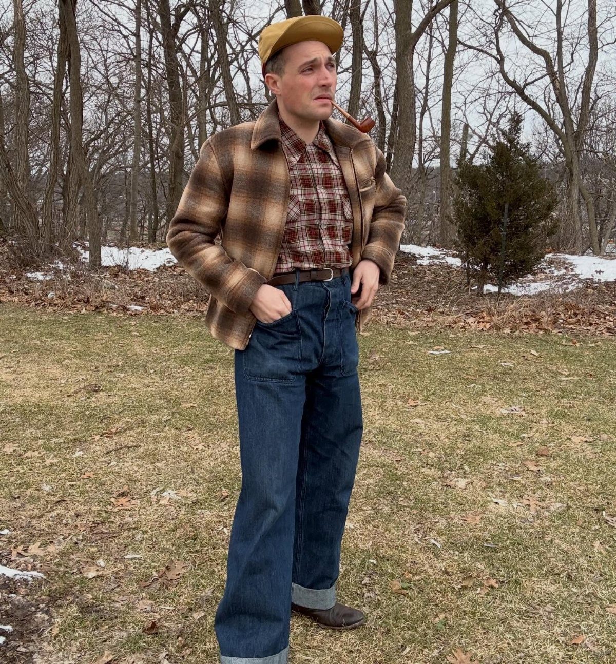 Man in plaid jacket, checkered shirt, and wide-leg denim jeans standing in a wooded field.