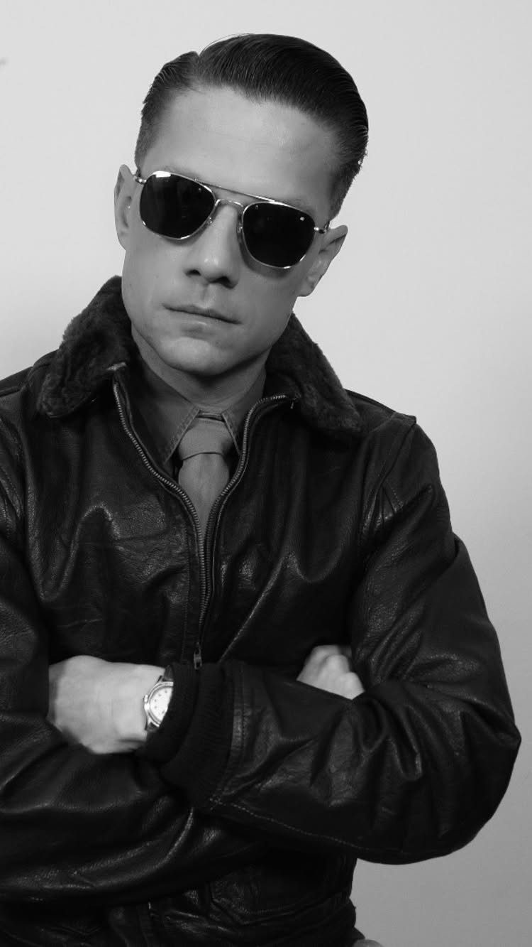 Man in aviator sunglasses and a leather jacket posing with arms crossed.