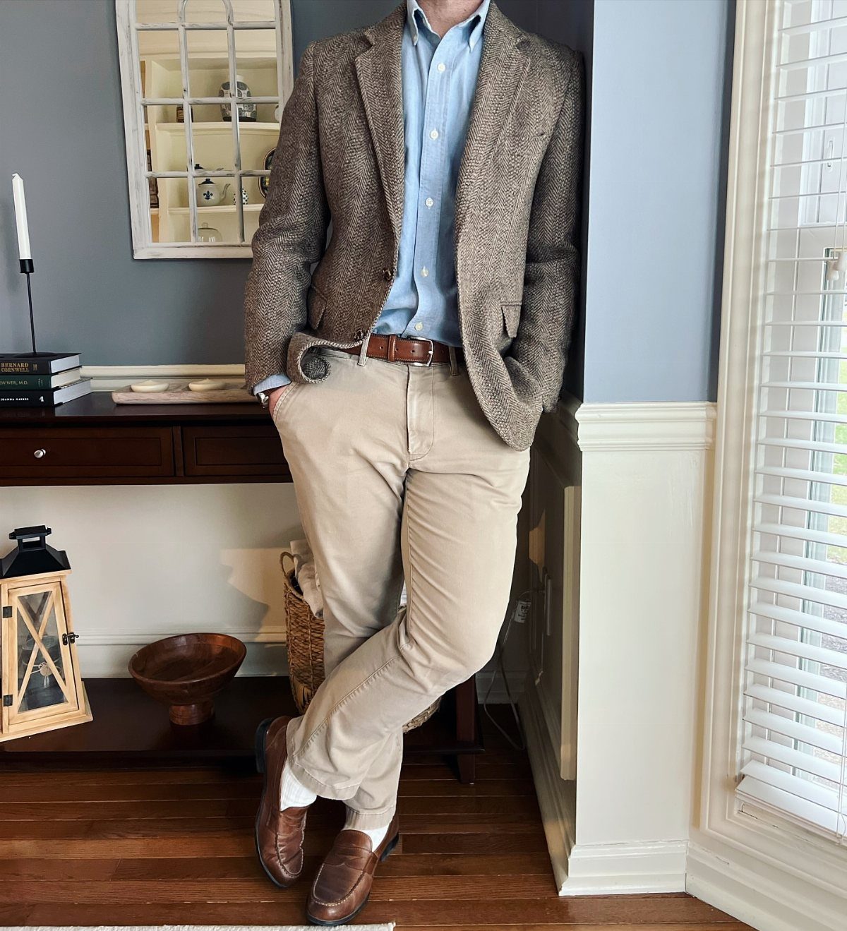 Man wearing a brown textured sport coat, light blue shirt, and khaki slacks leaning against a wall.