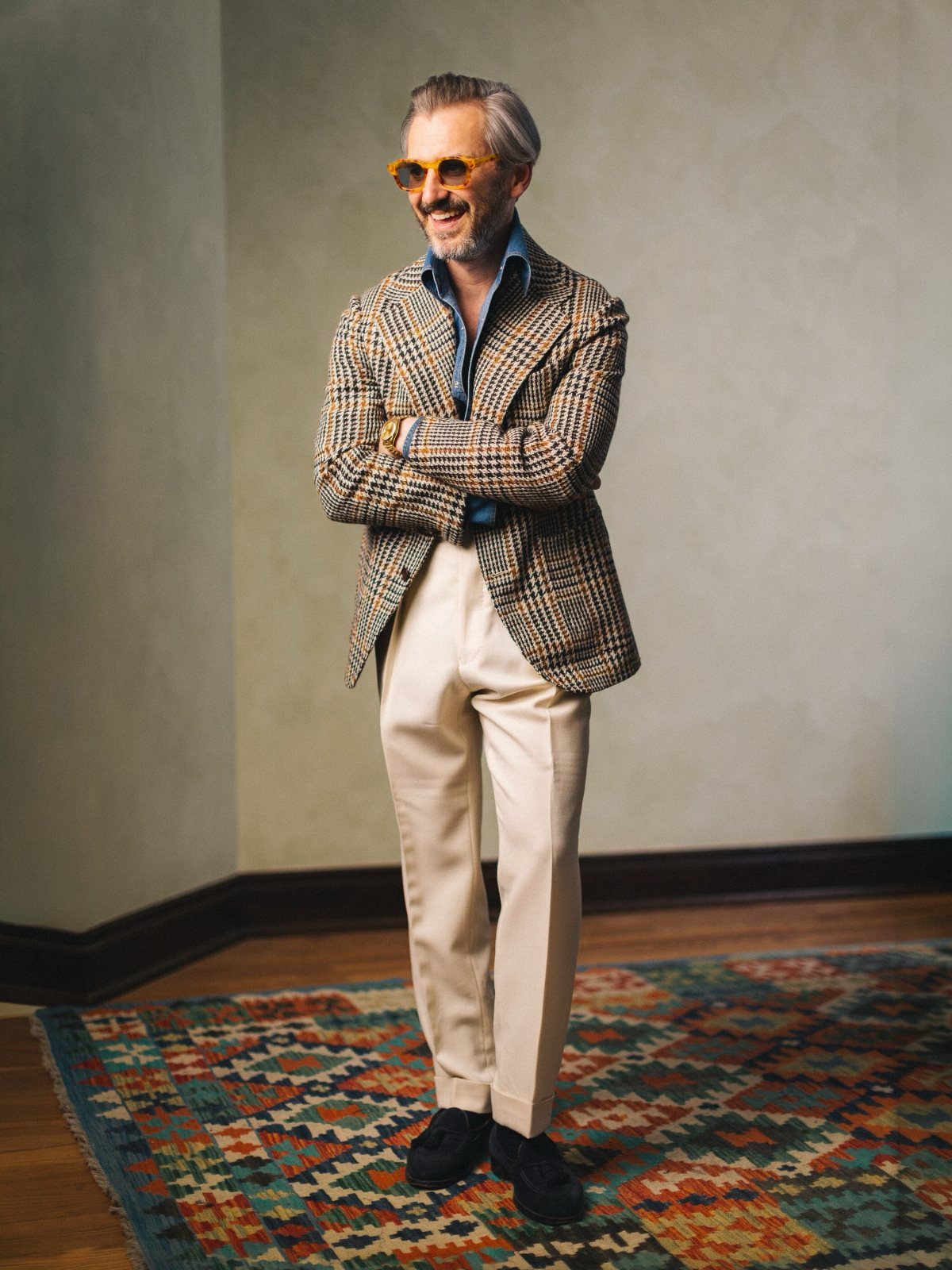 Smiling man in a patterned tweed blazer, denim shirt, and cream trousers standing on a rug.