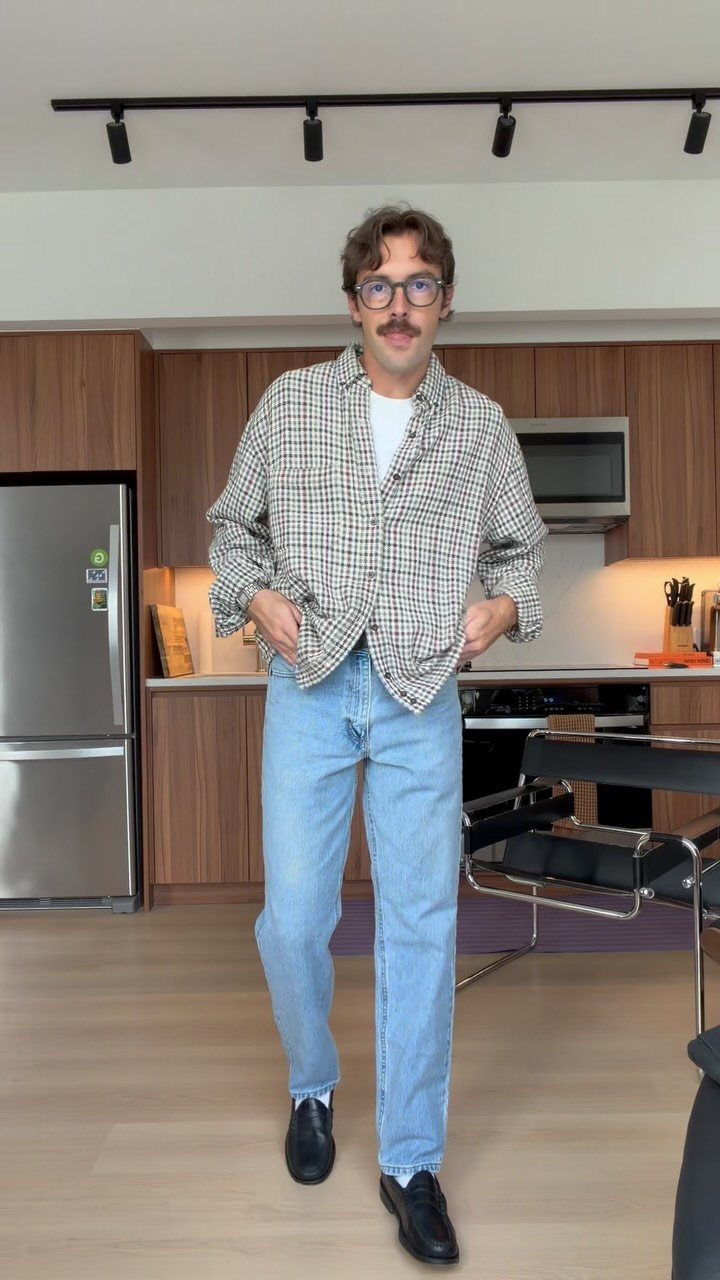 Man wearing a checkered button-down shirt, white t-shirt, and light blue jeans standing in a kitchen.