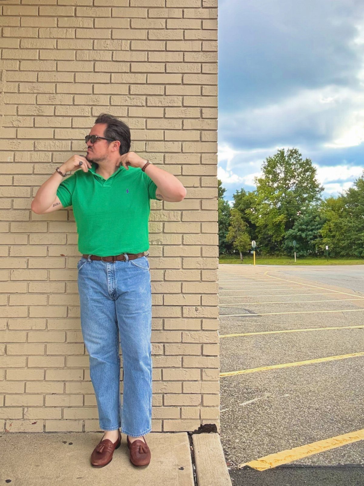 Man in a green polo and blue jeans standing against a brick wall.